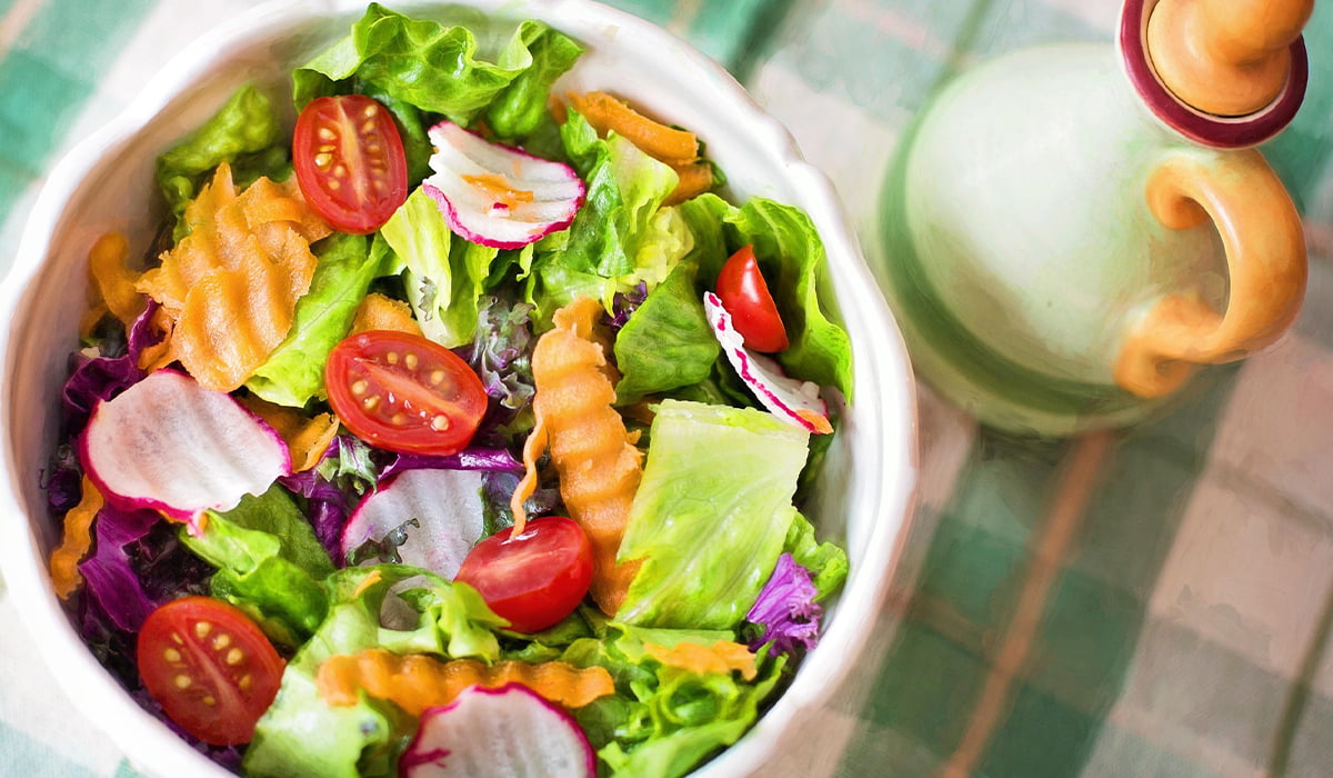 Insalatiera bianca con verdure miste fresche, lattuga, pomodorini e carote, accanto a un contenitore per condimenti